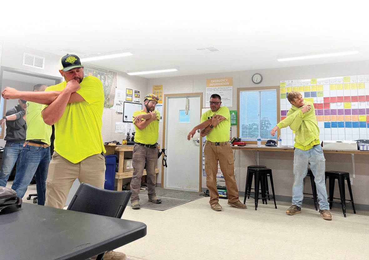 Construction crew performing shoulder stretches during a morning Stretch and Flex warm-up inside a jobsite trailer. Workers in high-visibility shirts prepare for the workday as part of C.D. Smith Construction’s safety routine.