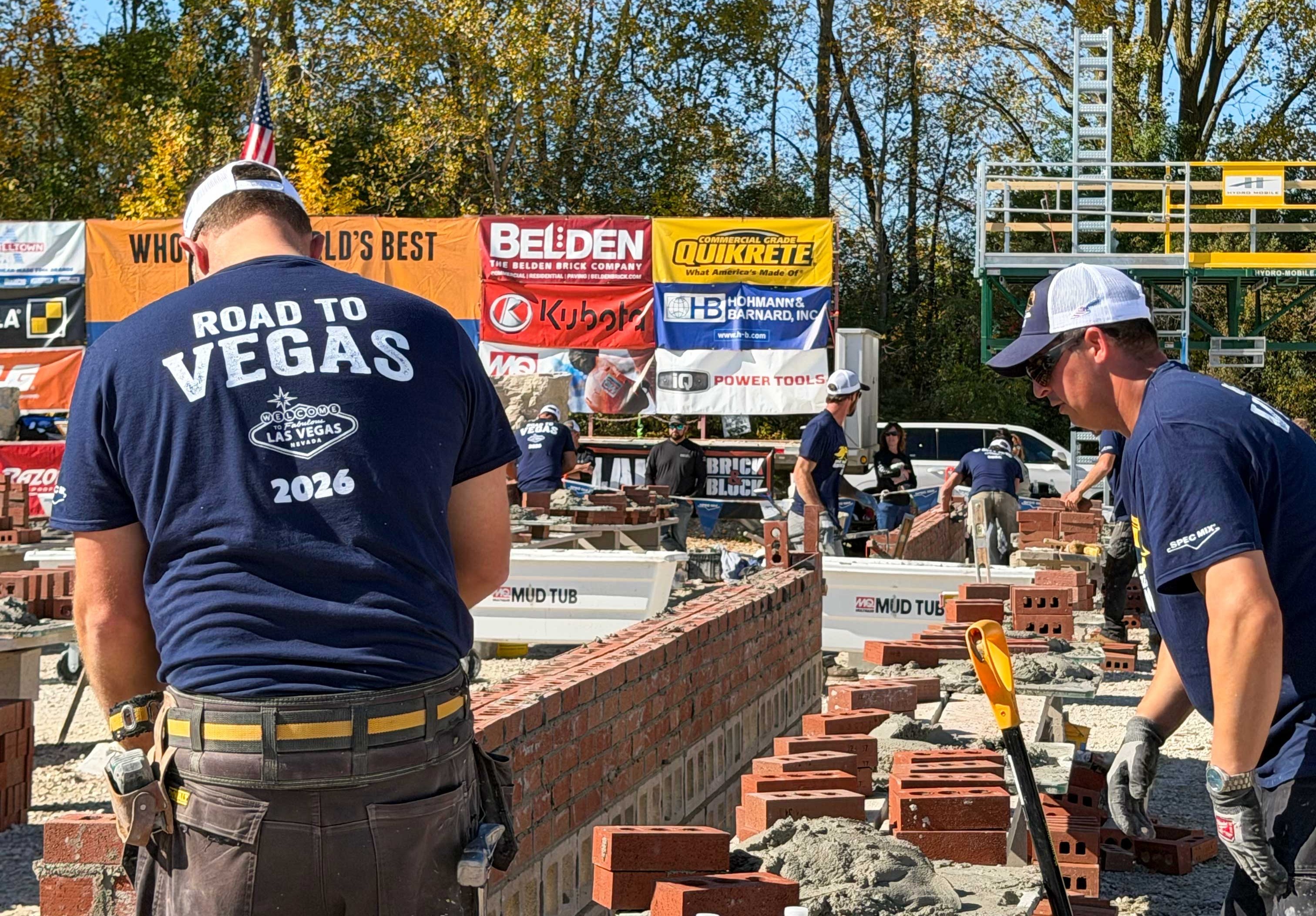 Masons work on competition walls during the 2026 SPEC MIX Bricklayer 500 Wisconsin Regional, with a Road to Vegas shirt in the foreground.