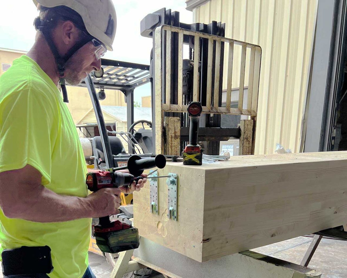 Construction worker installing Simpson Strong-Tie connectors on a glulam beam in a Wisconsin mass timber construction facility, preparing structural components for fire testing and high-rise building code compliance.