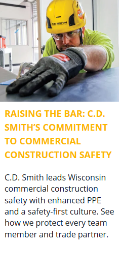 Focused construction worker wearing a CD Smith hard hat, safety glasses, and gloves while inspecting material alignment on a commercial interior project site in Wisconsin.