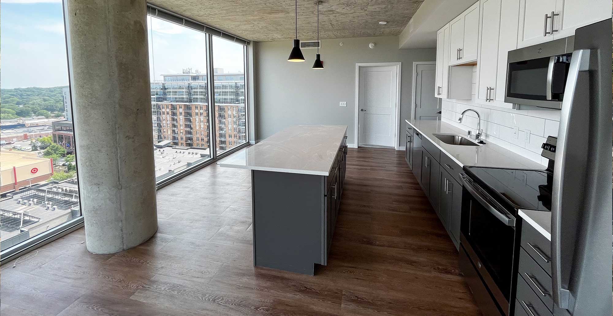 Open concept apartment kitchen at Madison Yards with large island, quartz countertops, stainless steel appliances, floor to ceiling windows, and exposed concrete column overlooking Madison skyline.