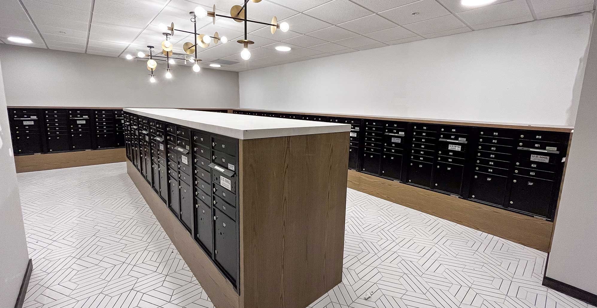Modern apartment building mailroom at Madison Yards featuring black resident mailboxes, wood wrapped cabinetry, white patterned tile flooring, and contemporary pendant lighting.