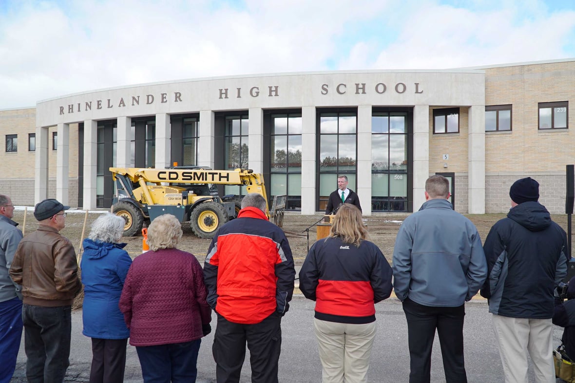 Speaker at podium at the Rhinelander High School groundbreaking ceremony, with staff and community members in attendance and CD Smith Construction equipment in the background.
