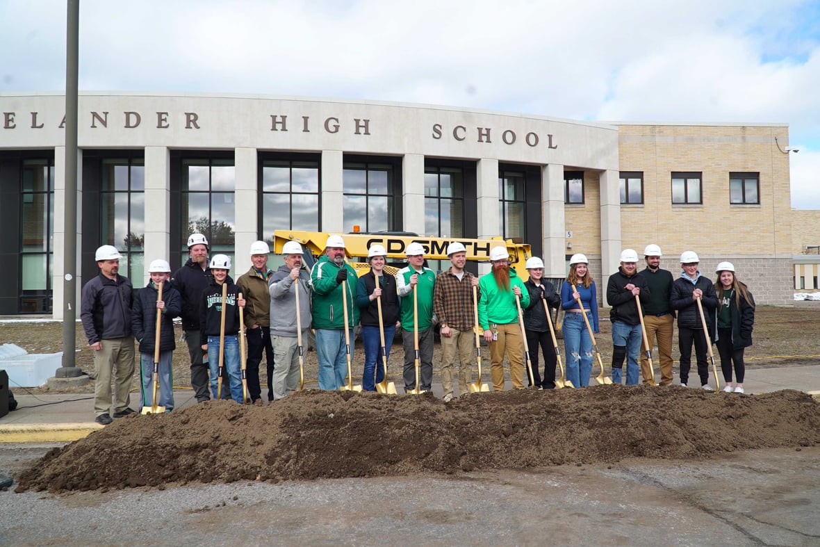 Group photo of students and staff smiling at the Rhinelander High School groundbreaking event, with CD Smith Construction equipment in the background.