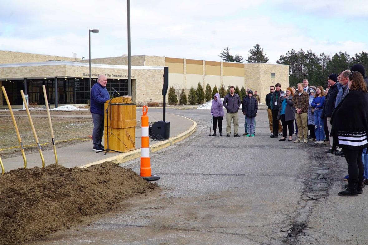 Superintendent Eric Burke speaking at the Rhinelander High School groundbreaking ceremony, standing at a podium with staff and community members in attendance.