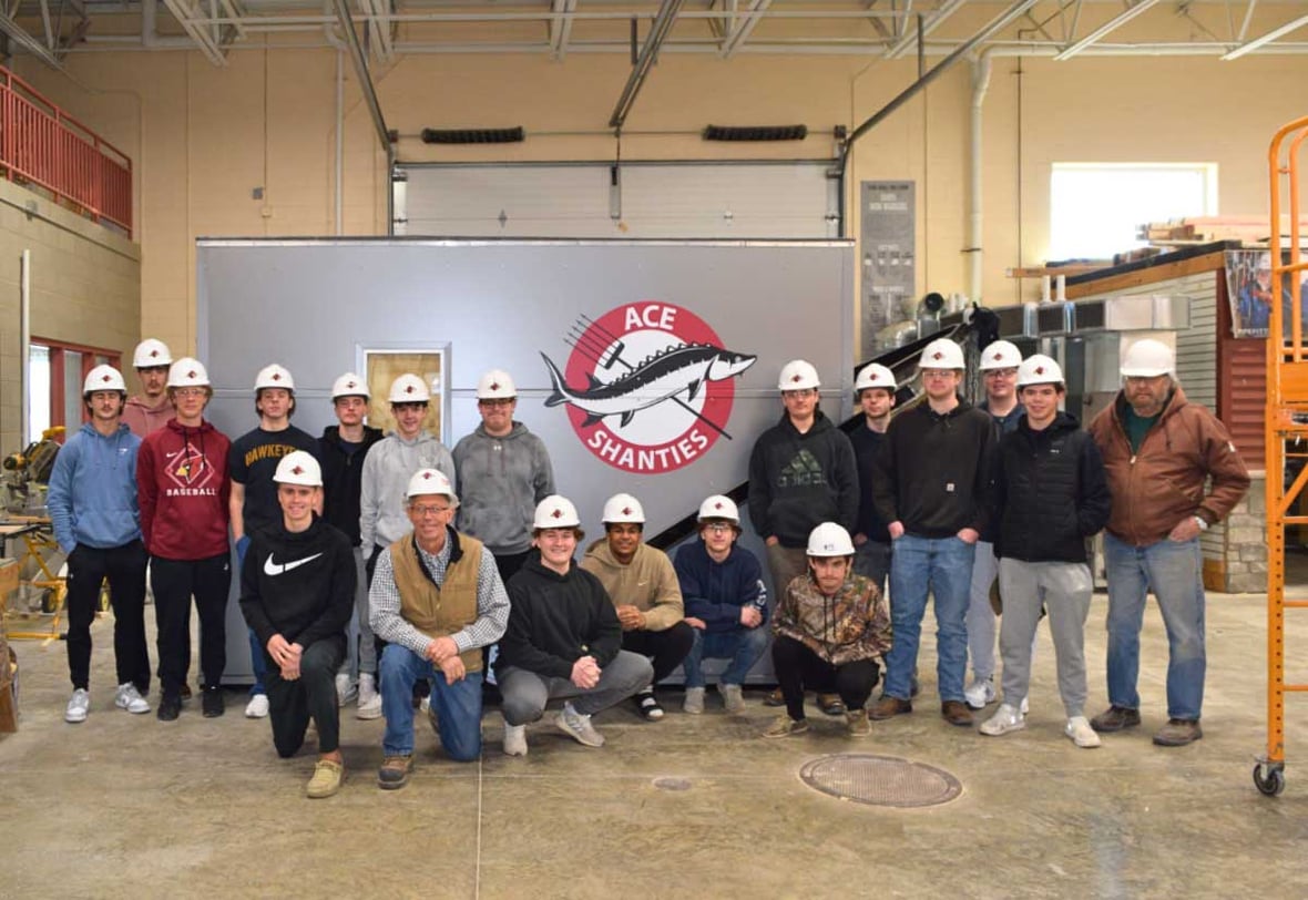 Fond du Lac High School ACE Academy students and local trades professionals stand together in a shop during the ACE Shanty construction project