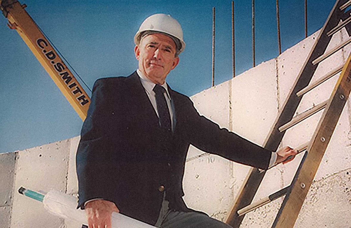 Tom Baker Sr. as President and CEO of C.D. Smith Construction on a jobsite in 1978 holding blueprints with a C.D. Smith crane visible in the background.