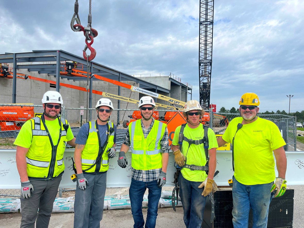 Construction workers in safety gear in front of a jobsite with a crane in the background ready to raise a beam at a commercial job site in Wisconsin, demonstrating proper PPE use with helmets, gloves, and high-visibility clothing.
