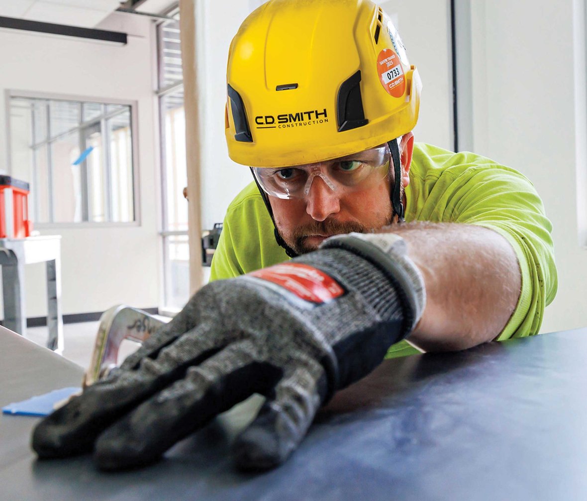 Focused construction worker wearing a CD Smith hard hat, safety glasses, and gloves while inspecting material alignment on a commercial interior project site in Wisconsin.
