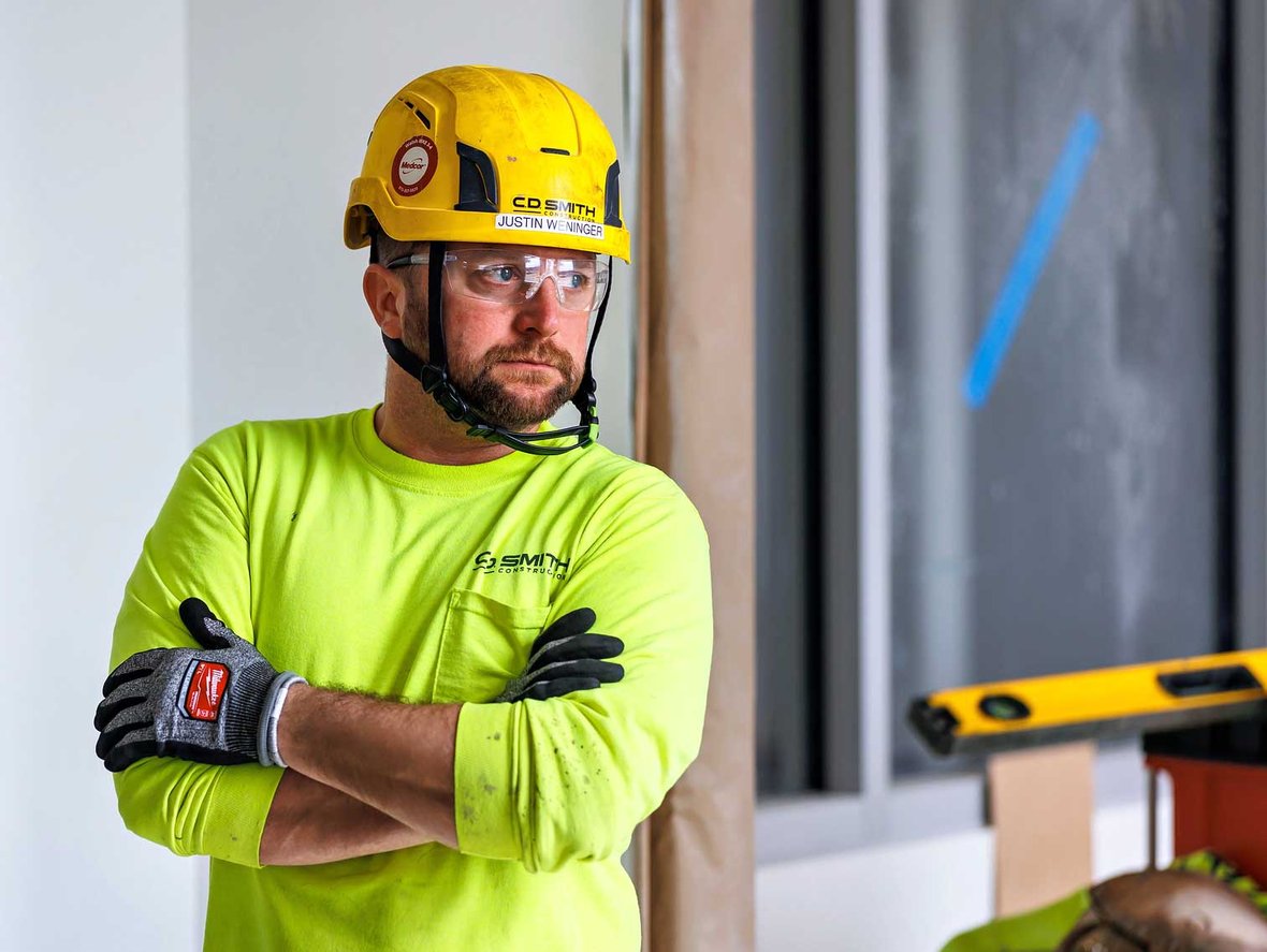 Focused construction worker wearing a CD Smith hard hat, safety glasses, and gloves while standing with arms crossed in a relaxed pose at a commercial interior project site in Wisconsin.