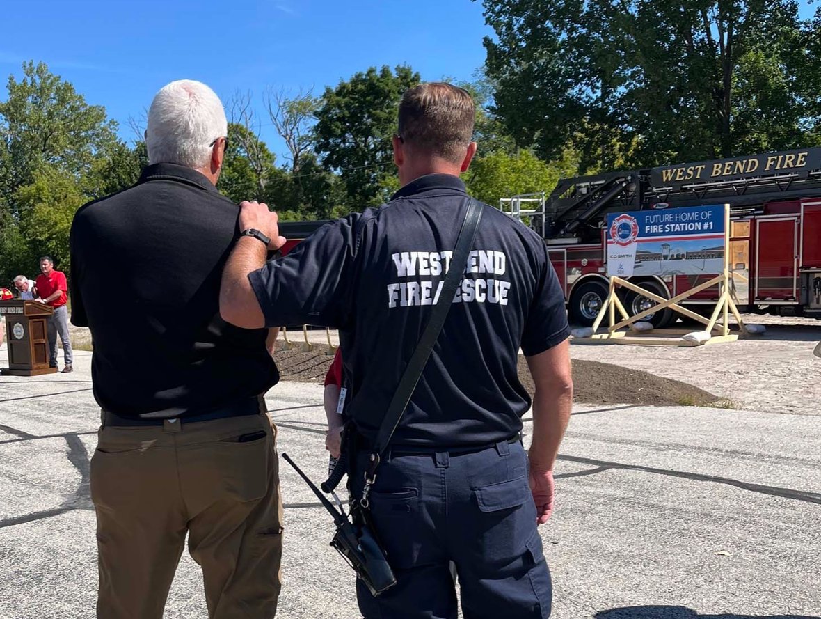 A firefighter rests a hand on a colleague’s shoulder while speakers present, with the project sign and ladder truck in view.