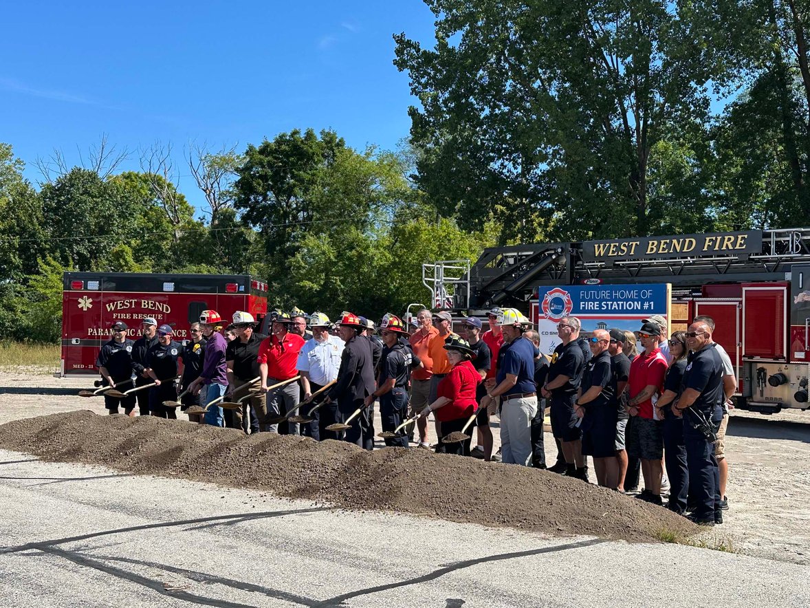Group of participants in hard hats and helmets holding shovels along the dirt mound at the Fire Station 1 groundbreaking